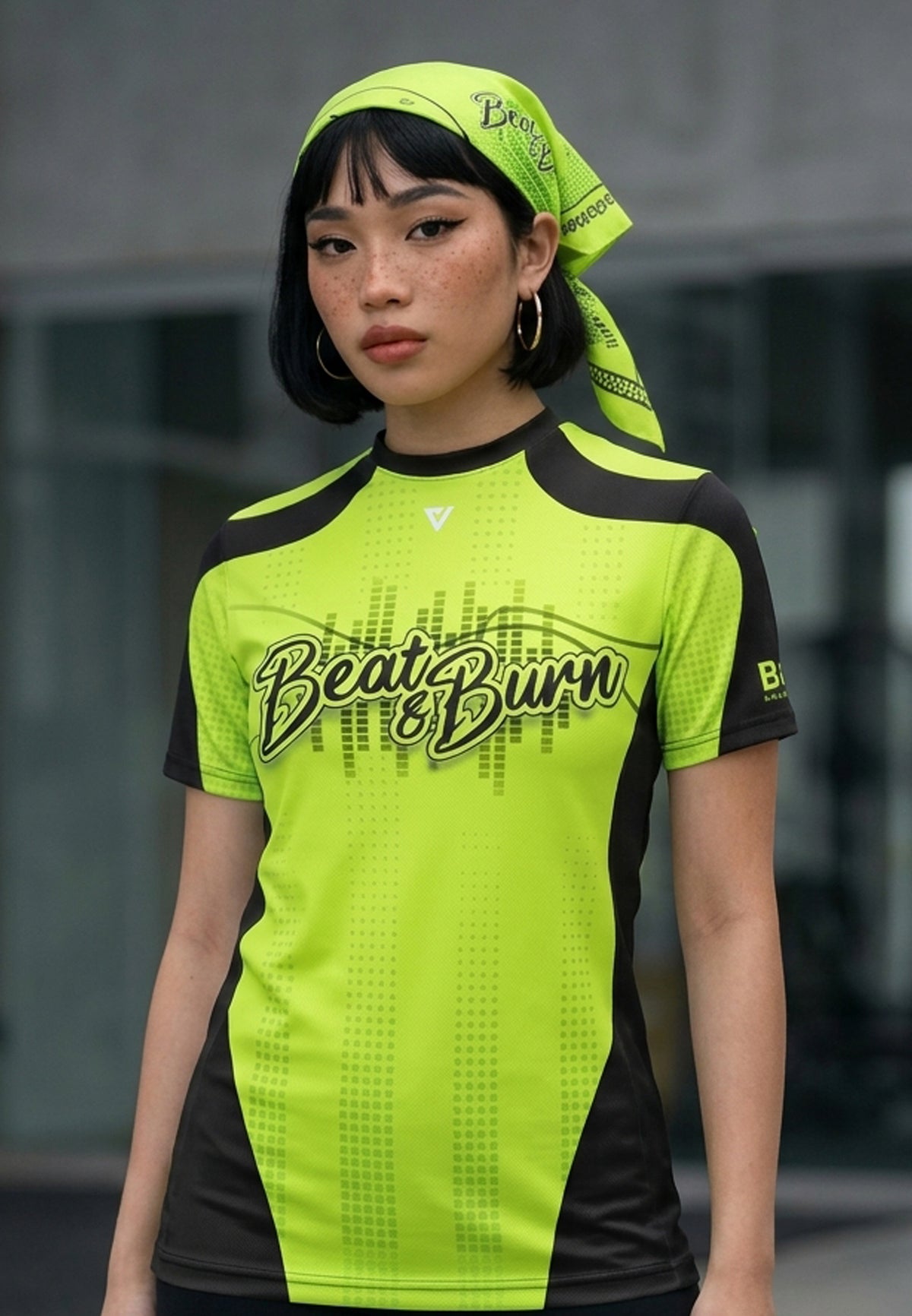 A woman wearing a bright neon yellow-green and black athletic t-shirt with 'Beat & Burn' printed across the chest, paired with a matching neon yellow-green headscarf, set against a blurred modern building background.