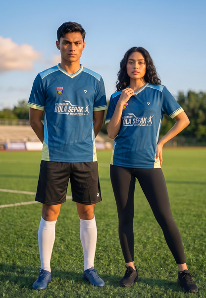 Two models standing on a grassy soccer field wearing matching blue and light blue V-neck athletic jerseys with 'Bola Sepak' printed on the front, paired with black shorts and leggings.